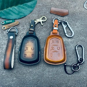 May include: Two leather key covers with keyrings. The key covers are brown and black with a textured leather finish. The keyrings are silver metal with a hook and a ring. The key covers have multiple buttons on the front. The keyrings have the text "SAM Luxury" and "BIG GOLDY" on them.