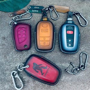 May include: Four leather car key fob covers with metal carabiner clips. One is deep magenta, one is gradient orange-brown, one is metallic blue-grey, and one is red with a black Sasquatch silhouette and text. Includes small personalized leather tags.
