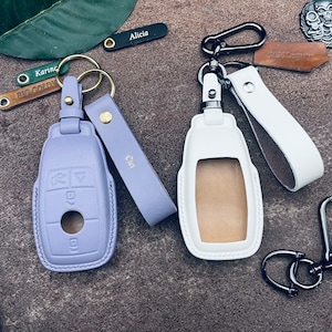 Op de afbeelding: Twee sleutelhangers met leren banden en sleutelhangers. De ene sleutelhanger is paars en de andere is wit. De paarse sleutelhanger heeft de naam "Chloe" op de band. De witte sleutelhanger heeft de naam "Sam Tracey" op de band.