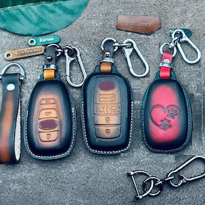 May include: A collection of leather key fobs and keychains. The key fobs are black and brown with silver hardware. One has a heart with paw prints, another has car button icons, and the third has a series of buttons. There are also two small tags with the names "Karina" and "Big Goldy".