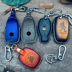 Op de afbeelding: Een verzameling leren sleutelhangers in verschillende kleuren, waaronder blauw, grijs en bruin. Sommige sleutelhangers hebben ontwerpen van autosleutels, terwijl andere unieke ontwerpen hebben, zoals een Sasquatch-silhouet en een Viking-symbool. Elke hanger heeft een metalen sluiting.