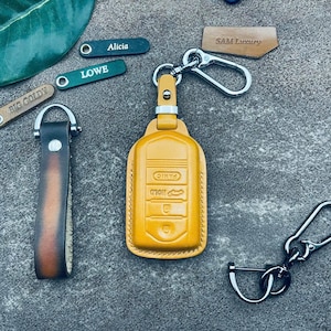 May include: A mustard yellow leather car key fob cover with embossed button icons including panic, boot, and lock symbols. It features a silver metal carabiner clip. Nearby are small leather tags labelled Alicia, LOWE, BIG GOLDY, and SAM Luxury.