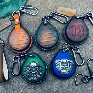 May include: A collection of leather keychains with metal clasps. Designs include an American flag, the number 13, and skull motifs. The keychains are in various colors, including brown, green, and blue. The text "SAM Luxury" is visible on a tag.