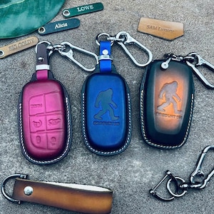 Könnte beinhalten: Drei Schlüsselanhänger-Etuis aus Leder in Magenta, Blau und einem braun-schwarzen Farbverlauf, jeweils mit Metallkarabiner. Die blauen und braunen Etuis zeigen eine Sasquatch-Silhouette und Text. Enthält kleine Lederanhänger mit Lowe, Alicia, Big Goldy und SAM Luxury.