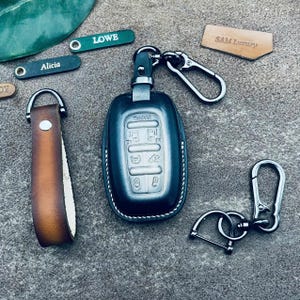 May include: A black leather key fob with a silver key chain and a silver key ring. The key fob has a silver button with the word "SHINKO" on it. The key fob is attached to a brown leather strap with a silver buckle. The key fob is on a grey surface.