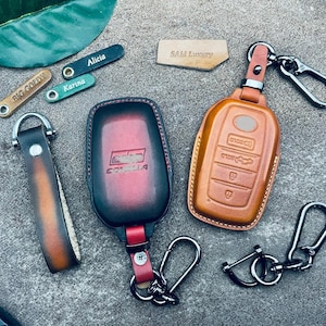 May include: Two leather keychains with key fobs. One keychain is brown with a black key fob that says "SAM Luxury". The other keychain is red and black with a key fob that says "COROLLA".