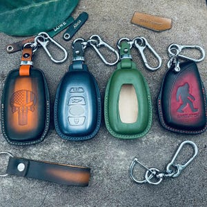 Puede incluir: Una colección de fundas de cuero para llaveros en varios colores, incluyendo naranja, azul y verde. Cada funda presenta un diseño único, como una calavera, una silueta de Sasquatch y un espacio en blanco. También se incluyen llaveros y etiquetas para llaves.