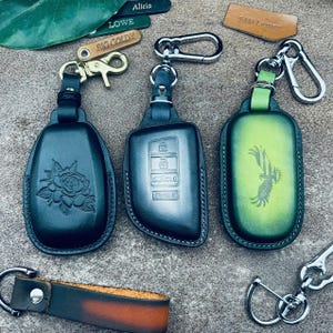 May include: Three leather car key fob covers with metal clips. One black cover features an embossed rose, one black cover shows button icons, and one green gradient cover displays an eagle graphic. Several small leather tags with names are visible.