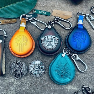 May include: Four leather keychains with different designs. The keychains are in the shape of a teardrop and have a metal ring attached. The designs include a skull with a motorcycle, a skull with an American flag, a skull with an American flag, and a rose. The keychains are on a gray surface.