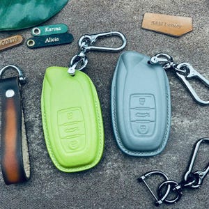 Könnte beinhalten: Eine Auswahl an Leder-Schlüsselanhängern in verschiedenen Farben. Enthält eine limettengrüne und eine graue Schlüsselanhängerhülle, einen braunen Lederriemen und personalisierte Anhänger mit Namen. Silberfarbene Hardware.