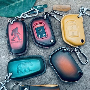 May include: A collection of leather key fob covers in various colors, including red, green, and yellow. Some covers feature embossed designs like a Sasquatch and a bear. Each cover has a metal keyring.