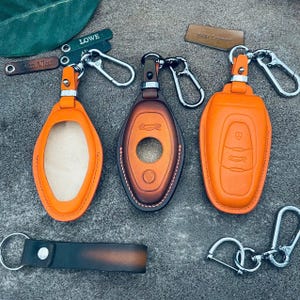 Könnte beinhalten: Drei Schlüsselanhänger-Etuis aus orangefarbenem und braunem Leder mit silbernen Schlüsselringen. Ein Etui ist offen, eines hat ein Autodesign und das dritte hat Autotasten. Ein passender Schlüsselanhänger aus Leder und zwei Schlüsselringe sind ebenfalls vorhanden.