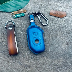 May include: A blue leather key fob with a silver key ring and a silver loop. The key fob has a small arrow pointing down and a round hole in the center. The key ring is attached to a brown leather strap with the text "SAM LEATHER" on it.