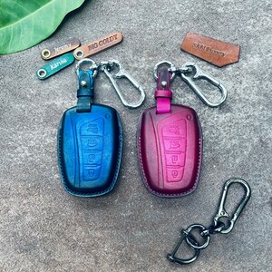 May include: Two leather key covers, one blue and one burgundy, with silver keychains and a silver swivel clip. The key covers have four buttons each and are labeled "BIG GOLDY" and "SAM Luxury".