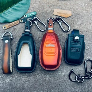 Peut inclure: Trois porte-clés en cuir avec des designs différents. Le porte-clés à gauche est noir avec une découpe pour une clé. Le porte-clés du milieu est orange et rouge avec un design qui ressemble à une clé de voiture. Le porte-clés à droite est noir avec un design qui ressemble à une clé de voiture. Les porte-clés sont tous attachés à des anneaux métalliques.