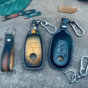 Op de afbeelding: Leren sleutelhangers en sleuteletuis in verschillende stijlen. De sleuteletuis zijn zwart en bruin met zilverkleurige hardware. Eén sleutelhanger heeft een leren bandje met een knoop. Twee kleine groene sleutelhangers hebben de namen "Alicia" en "Lowe" gegraveerd.