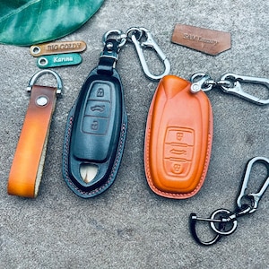 May include: Two leather keychains with key fobs. One keychain is black with red stitching and the other is orange with brown stitching. Both keychains have a silver metal key ring and a silver metal hook.