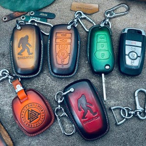 May include: A collection of leather key fob covers in various colors, including brown, green, and red. Some covers feature a Sasquatch silhouette, while others display car key designs. Keychains and metal clasps are attached.