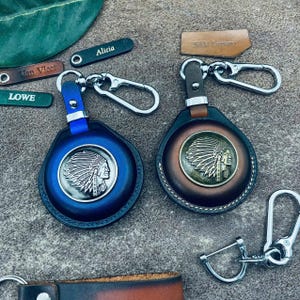 May include: Two leather keychains with metal accents. One is blue, the other is brown. Each features a Native American profile in a headdress. Additional small leather tags with names are also visible.