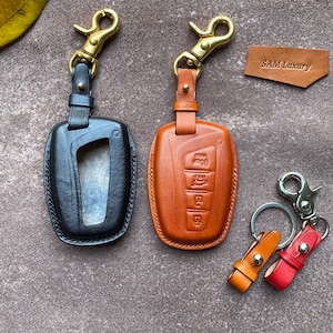 May include: Two leather keychains with metal key rings. One keychain is black and has a key fob slot. The other keychain is brown and has a design of buttons. The text "SAM Luxury" is visible on a brown tag.