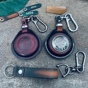 Può includere: Due custodie rotonde per chiavi in pelle con clip in metallo, una con un design in rilievo di copricapo nativo americano e l'altra con un emblema in metallo argentato dello stesso motivo. Include un portachiavi in pelle rettangolare e targhette.