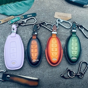 Op de afbeelding: Vier leren sleutelhangers met verschillende gekleurde sleutelhoezen. De sleutelhoezen zijn paars, zwart, oranje en groen. De sleutelhangers zijn bevestigd aan een zilveren metalen ring. De sleutelhangers zijn gelabeld met de namen "Big Goldy", "Karina", "Alicia" en "SAM Luxury".