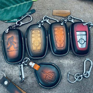 May include: A collection of leather key fobs in various colors, including black, brown, and red. Some fobs feature embossed designs like a Sasquatch and a hand gesture. Each fob is attached to a silver keychain.