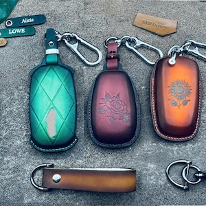 May include: Three leather keychains with different designs. The keychain on the left has a green diamond pattern, the middle keychain has a rose design, and the keychain on the right has a sunflower design. Each keychain has a silver metal ring and a silver metal hook.