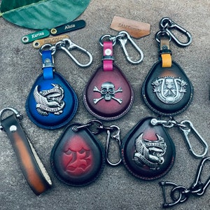 Pode incluir: Seis chaveiros de couro com designs diferentes. Os chaveiros são de várias cores, incluindo azul, vermelho, preto e marrom. Cada chaveiro apresenta um design diferente, incluindo uma caveira e ossos cruzados, um pássaro com as asas abertas, uma ferradura e um número 23. Os chaveiros estão todos presos a argolas de metal prateadas.