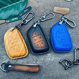 May include: Three leather keychains with different colored key covers. The key covers are brown, black, and blue. The keychains are attached to a silver metal ring. The key covers have a design of buttons on them. The keychains are labeled with the words "LOWE", "Alicia", and "SAM Luxury".