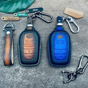 May include: Two leather keychains with key fobs. One keychain is black with orange accents and the other is blue. Both keychains have a metal ring and a metal hook. The keychains are labeled with the names "Alicia" and "SAM Luxury".