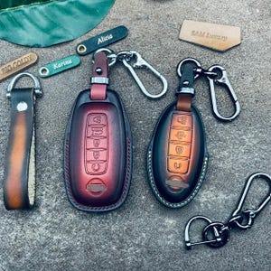 May include: Two leather keychains with key fobs. The keychains are brown and black with a red and orange gradient. The key fobs are black and silver. The keychains are labeled "SAM Luxury" and "Alicia".