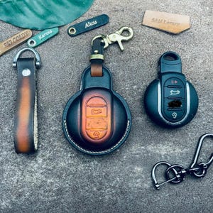 以下が含まれることがあります： ブラウンとブラックのグラデーション仕上げが施された円形のレザー製カーキーフォブカバー。ゴールドトーンの金属製スイベルクラスプ付き。お揃いのレザーストラップ、黒いキーフォブ、BIG GOLDY、Karina、Alicia、SAM Luxuryと刻印された小さなレザータグが含まれています。