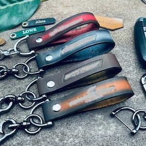 May include: Four leather keychains with metal hardware, each featuring a loop design secured by a silver rivet. The colors include a red and black gradient, a solid dark blue, a dark brown, and a tan and black gradient, all with metal clips.