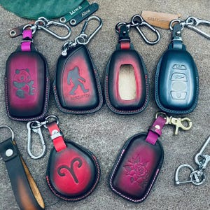 Puede incluir: Una colección de fundas de cuero para llaveros en varios colores, con diseños como un panda, un Bigfoot y una rosa. Cada funda tiene un llavero de metal y una correa en contraste. Los llaveros son en tonos rojos, negros y morados.