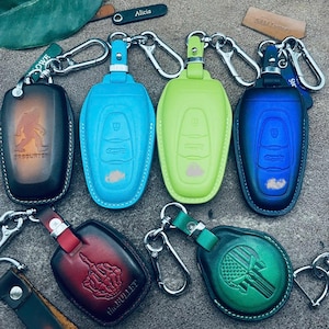 Könnte beinhalten: Eine Sammlung von sechs Leder-Schlüsselanhängern mit verschiedenen Designs. Die Schlüsselanhänger sind in verschiedenen Farben, darunter Braun, Blau, Grün, Rot und Hellblau. Die Schlüsselanhänger zeigen verschiedene Designs, darunter einen Schädel, einen Mittelfinger, einen Bären und eine Flagge. Die Schlüsselanhänger sind an Metallschlüsselringen befestigt.