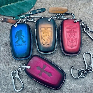 May include: Four rectangular leather key fob covers with metal carabiner clips. One blue cover features a Sasquatch silhouette and text, one brown cover shows button icons, one red cover displays button icons, and one maroon cover has a cross design.