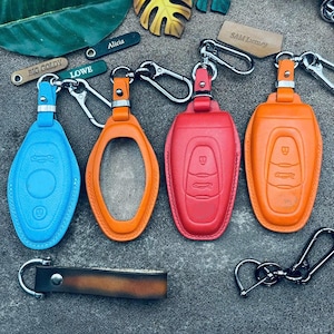 Könnte beinhalten: Eine Auswahl an Leder-Schlüsselanhängern in Blau, Orange und Rot. Einige haben ein Autodesign, andere sind offen oder haben einen Lederriemen. Die Schlüsselanhänger sind an silbernen Karabinerhaken befestigt. Maße in cm.