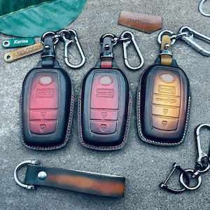 May include: Three leather key covers with different colored designs. The key covers are attached to key rings with metal clasps. The key covers are labeled "Karina", "BIG GOLDY", and "SAM Luxury".