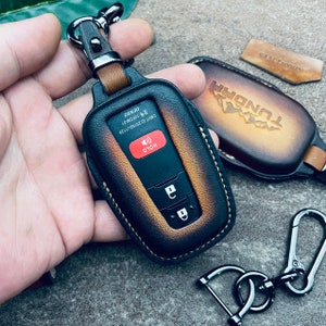 May include: A brown leather key fob with a black leather key cover. The key cover has a red button labeled "HOLD" and a silver key chain with a silver carabiner. The key fob has the word "TUNDRA" embossed on it.