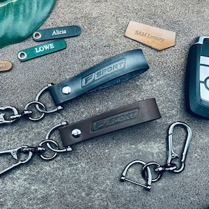 May include: Two leather keychains with silver metal rings and a silver carabiner clip. The keychains are brown and black and have the text "F SPORT" embossed on them.