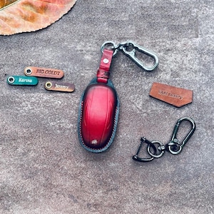 Peut inclure: Porte-clés en cuir rouge avec un anneau de clé argenté et une sangle en cuir noir. Le porte-clés a un design cousu et est attaché à un anneau de clé argenté avec une sangle en cuir noir. Le porte-clés est étiqueté "BIG GOLDY" et "SAM Luxury".