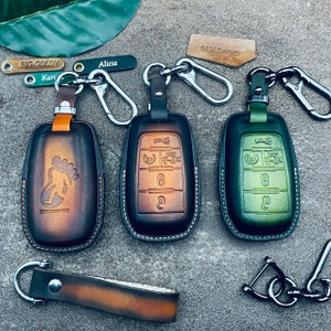 May include: Three leather key fob covers with metal clips, featuring a brown Bigfoot design, a dark brown cover, and a green cover. Small leather tags are included with the text BIG GOLDY, Alicia, Karin, and SAM Luxury. The covers have embossed button icons.