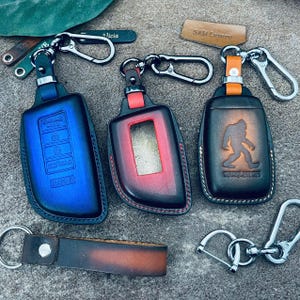 May include: Three leather car key fob covers with metal clips. One is blue, one is red and black, and one is brown with a Bigfoot graphic and the text "SASQUATCH". Includes a brown leather keychain strap and two small leather tags labeled "Alicia" and "SAM Luxury".