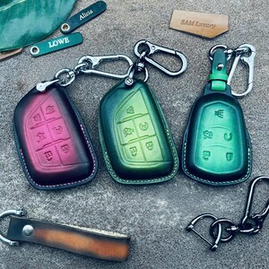 May include: Three leather key covers with different colored designs. The key covers are green, pink, and green. The key covers have a stitched edge and are attached to key rings. The key rings are attached to a leather strap with the text "SAM Luxury" on it.