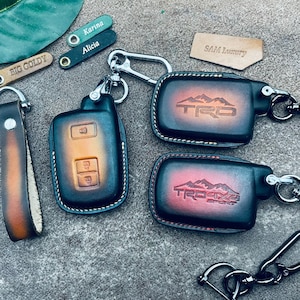 May include: Four leather keychains with different designs. The keychains are brown and black with red and yellow accents. The keychains feature the text "BIG GOLDY", "Karina", "Alicia", "SAM Luxury", and "TRD".