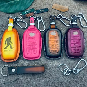 Peut inclure: Plusieurs étuis de clés en cuir de différentes couleurs, dont orange, rose et bordeaux. Certains étuis présentent des motifs en relief et le mot "SASQUATCH". Des porte-clés et de petites étiquettes en cuir avec des noms sont également visibles.