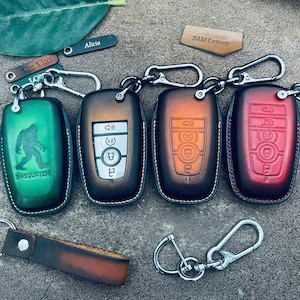 May include: A collection of leather key fob covers in various colors, including green, brown, orange, and red. The green fob features a Sasquatch graphic. Each fob has a silver carabiner clip for attachment.