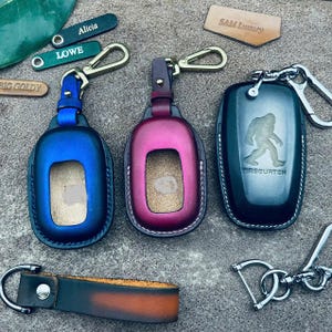 May include: Three leather keychains with different colored key covers. The key covers are blue, red, and black. The black key cover has a graphic of a sasquatch and the text "Sasquatch".