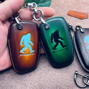 May include: Three leather key fob covers featuring a Sasquatch silhouette and the text SASQUATCH. The covers are brown, green, and dark brown, each with a silver metal ring attachment, visible stitching along the edges, and a smooth finish.